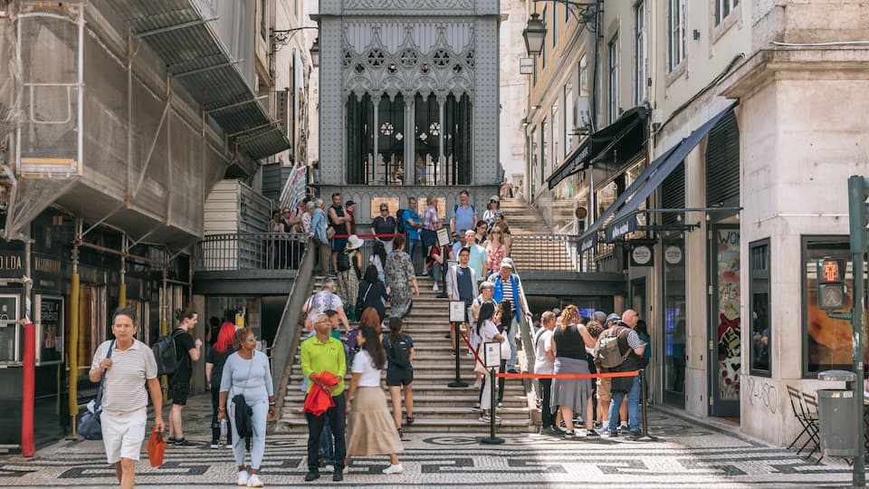 Una vivace strada cittadina con una folla che sale una scalinata verso un edificio con finestre in stile gotico finemente decorate.