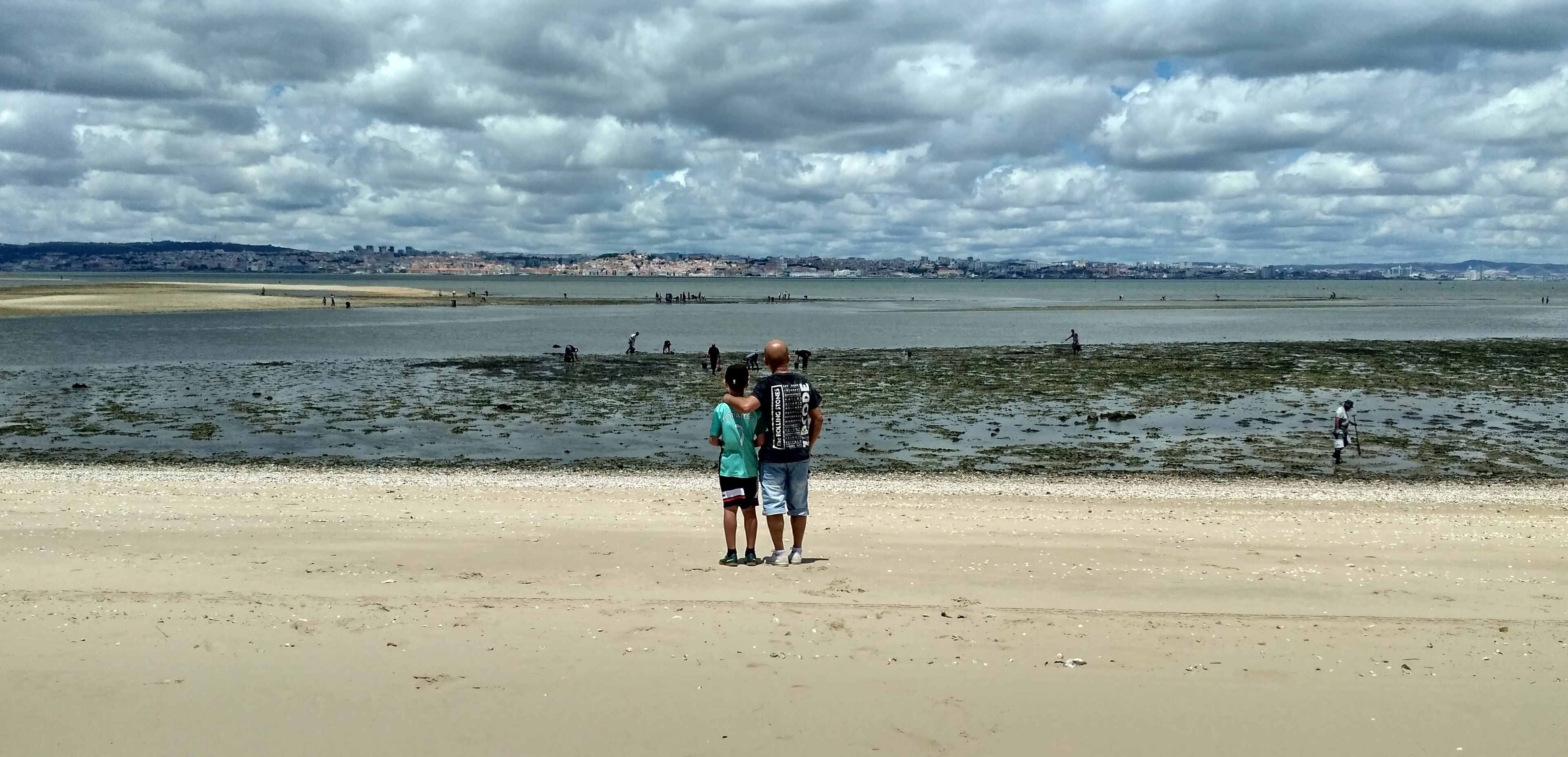 A beach with people standing on the sand, a large body of water, and a distant city skyline under a cloudy sky.