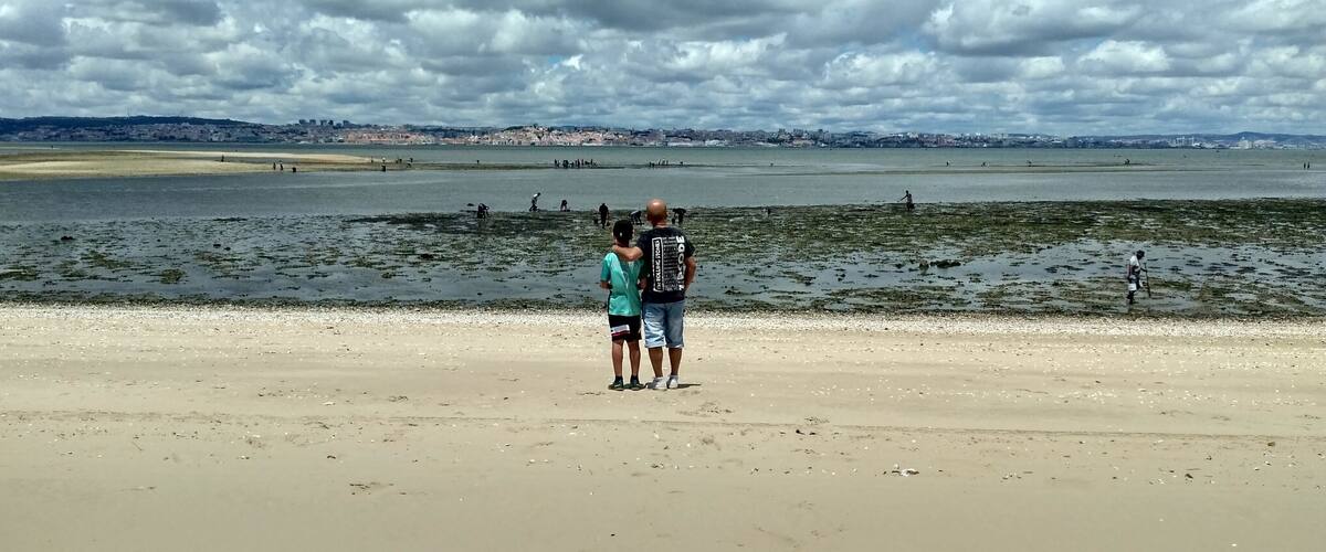 A beach with people standing on the sand, a large body of water, and a distant city skyline under a cloudy sky.