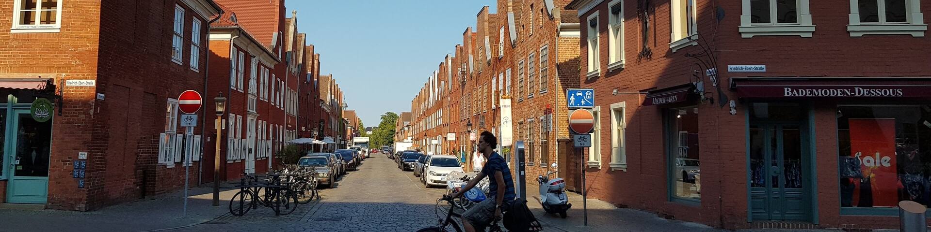 A cobblestone street lined with red brick buildings, a cyclist, parked cars, and a 'sale' sign on a storefront.
