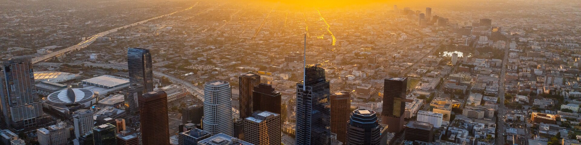 Paisagem urbana com um arranha-céu imponente, um estádio grande e uma área urbana densa ao pôr do sol.