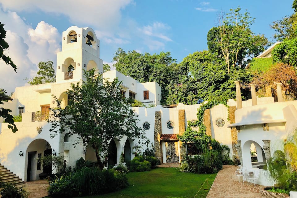 A white colonial-style building with arched doorways and a bell tower, surrounded by lush greenery and a brick pathway.