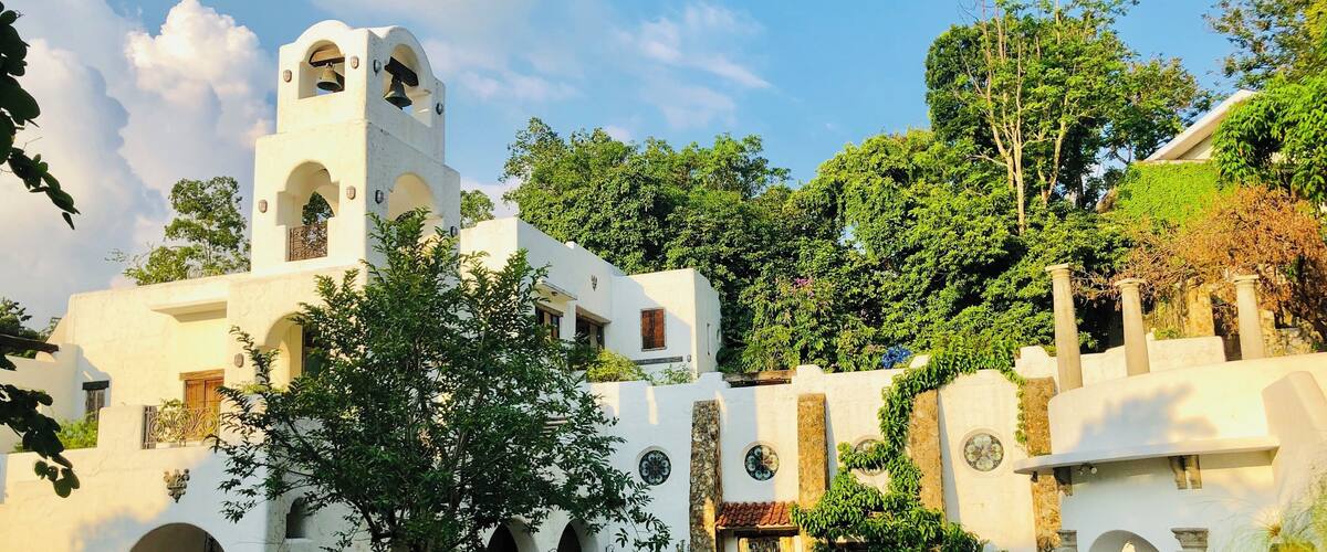 A white colonial-style building with arched doorways and a bell tower, surrounded by lush greenery and a brick pathway.