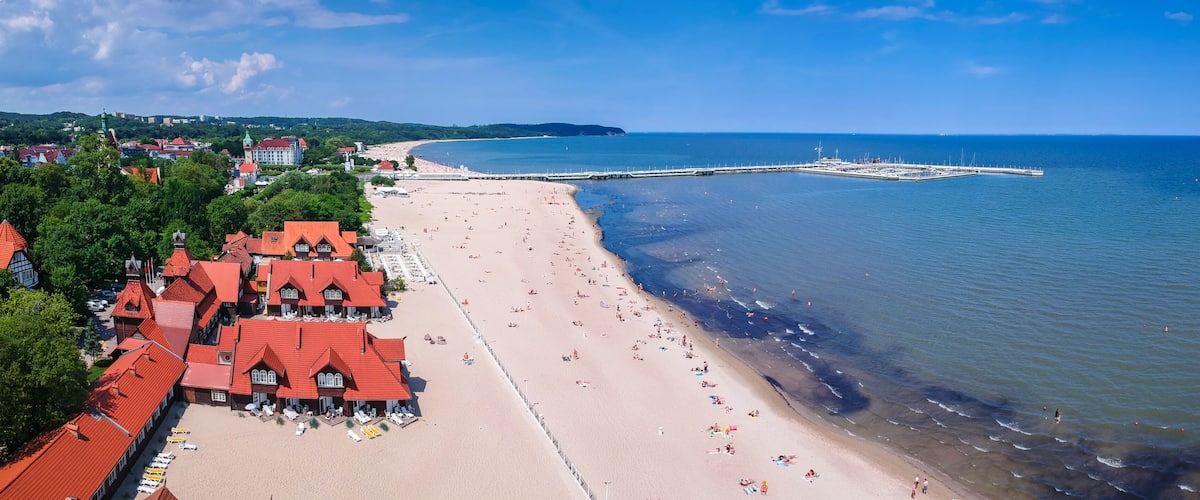 A coastal town with a sandy beach, red-roofed buildings, and a pier extending into the sea.