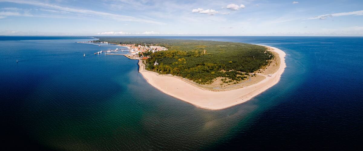 An aerial view of a coastal island with a sandy beach, dense greenery, and a clear blue sea.