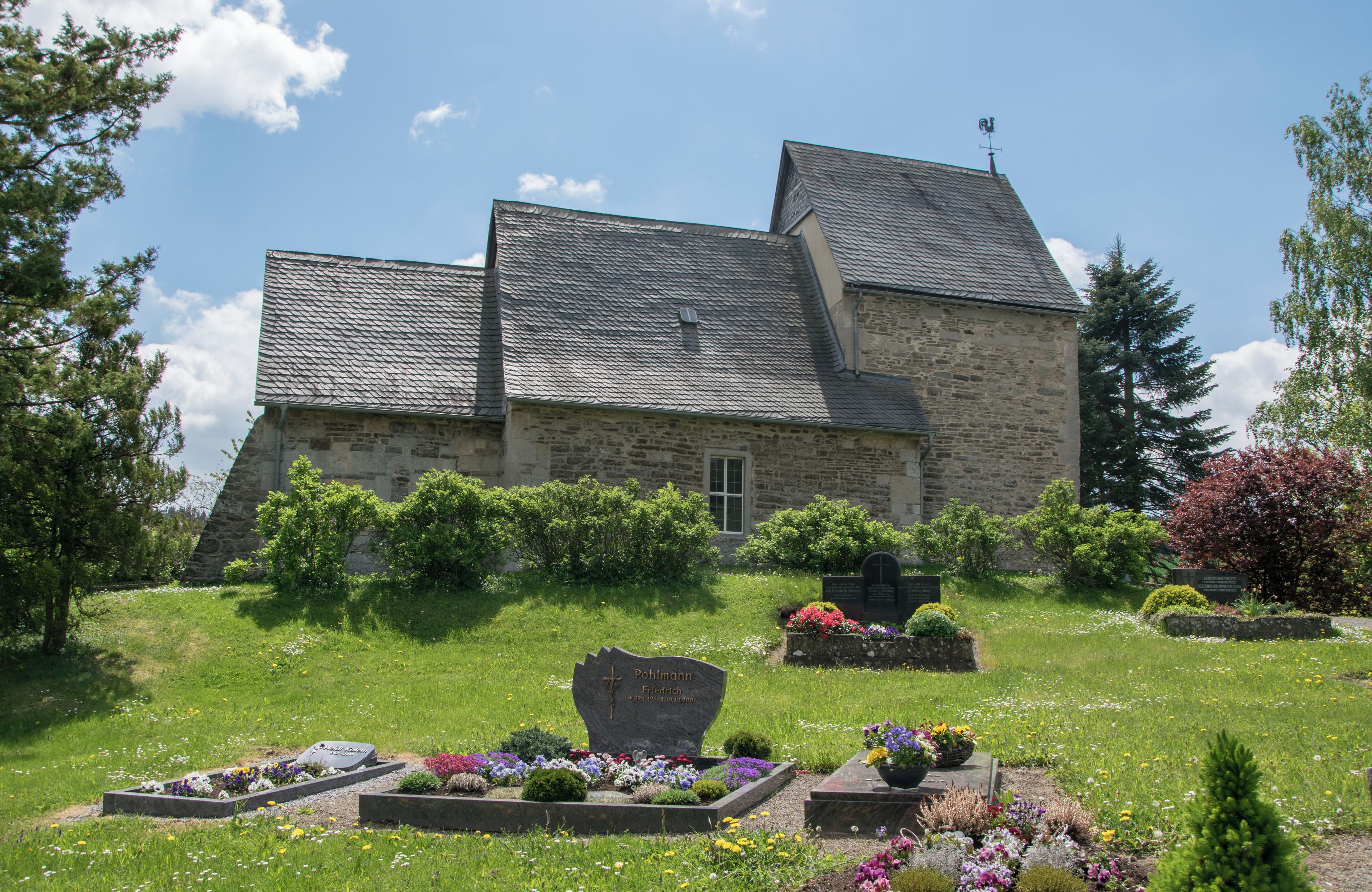 A stone building with a sloped roof, surrounded by a well-maintained garden with various flowers and shrubs.
