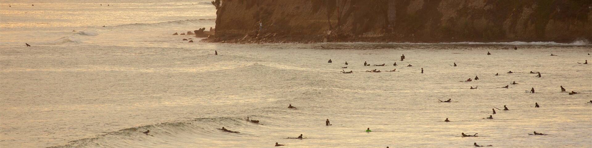 A coastal scene with surfers in the water, a sunset, and buildings on a cliff.