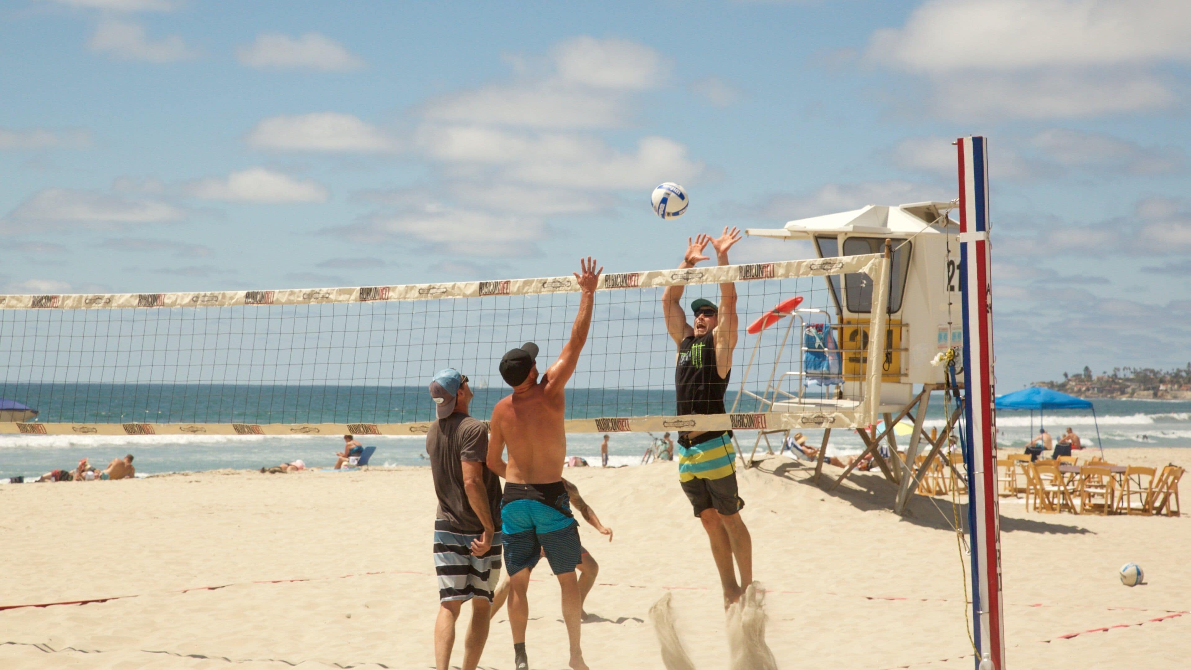 A beach volleyball game with players, a lifeguard tower, and the ocean in the background.