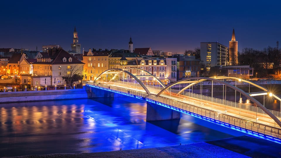 A nighttime cityscape with a brightly lit bridge over a river, historic buildings, and a prominent tower in the background.