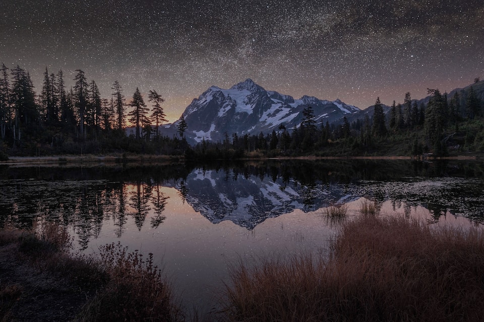 A mountain range reflected in a calm lake at night with a starry sky.