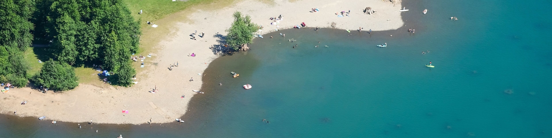 Eine Vogelperspektive eines Strandes mit klarem Wasser, umgeben von dichtem Wald, mit mehreren Booten und Personen.