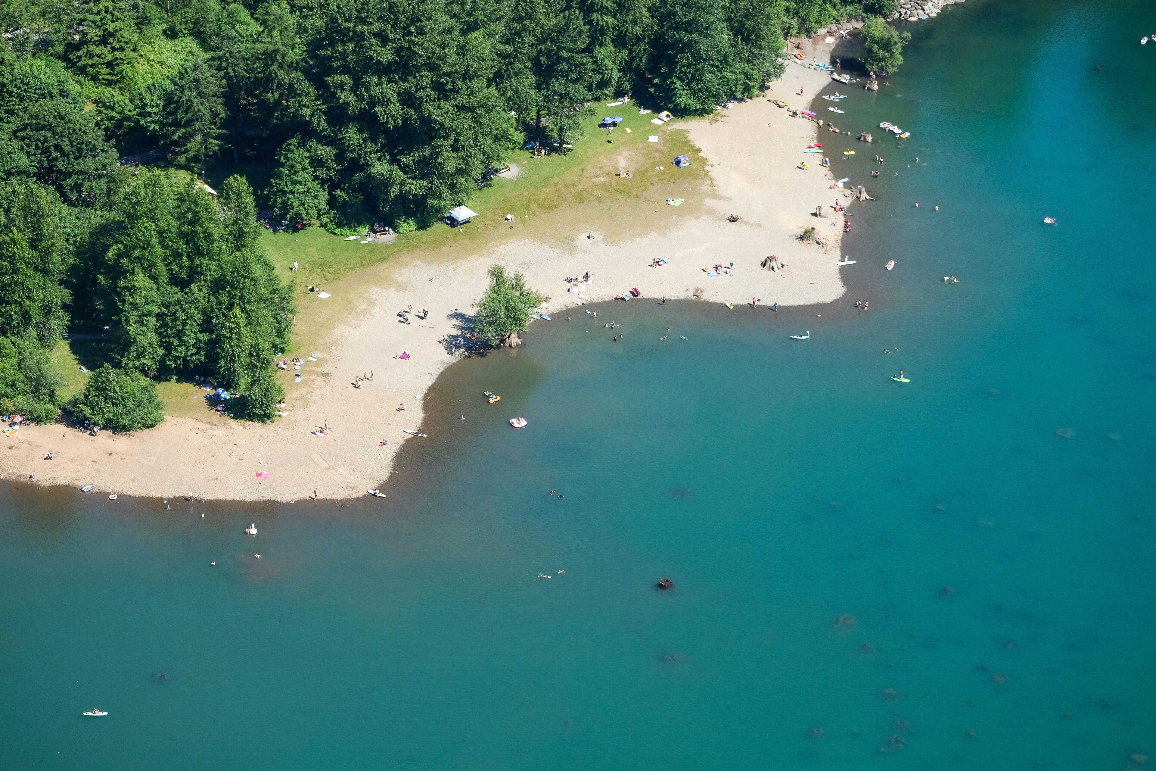 An aerial view of a beach with clear waters, surrounded by dense forest, and several boats and people visible.