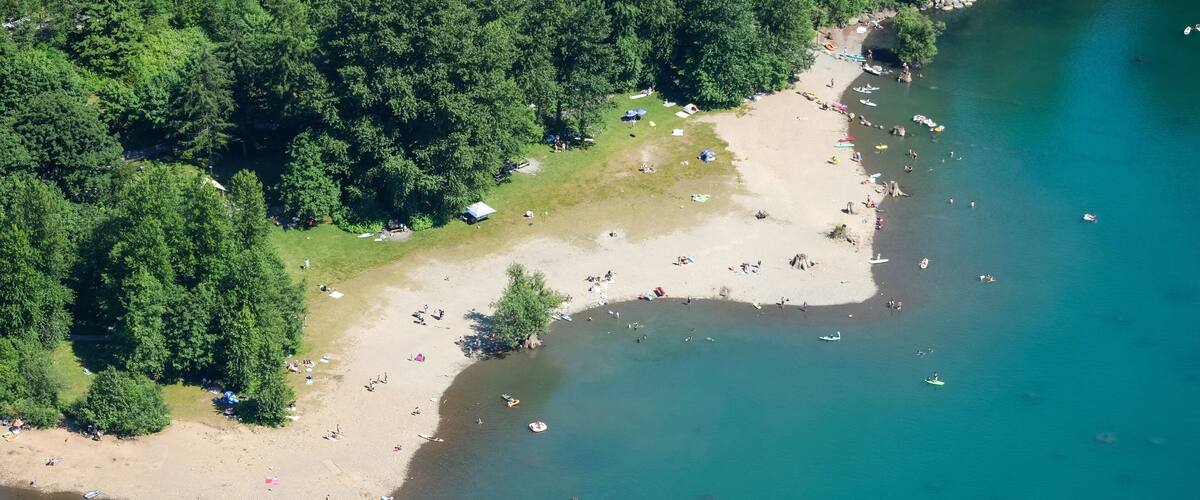 An aerial view of a beach with clear waters, surrounded by dense forest, and several boats and people visible.