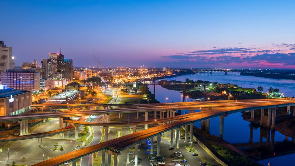 A cityscape at dusk with illuminated buildings, a river, and a bridge.