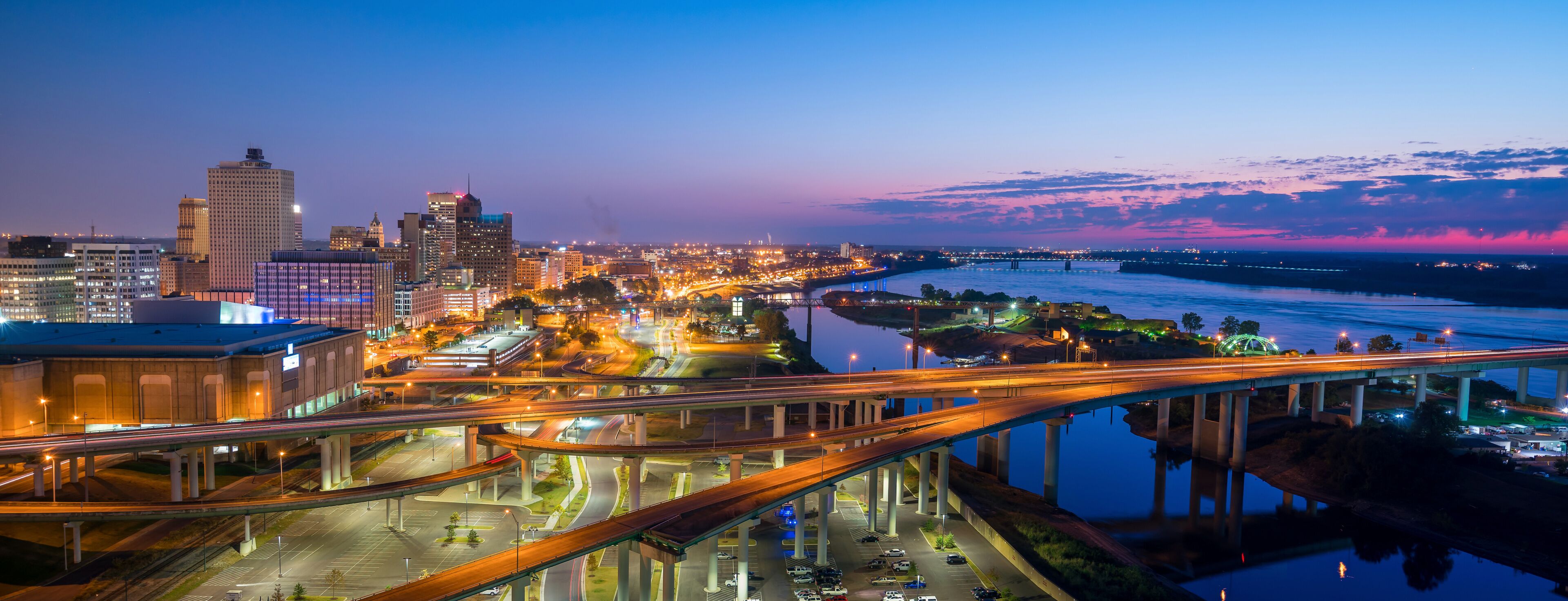 A cityscape at dusk with illuminated buildings, a river, and a bridge.