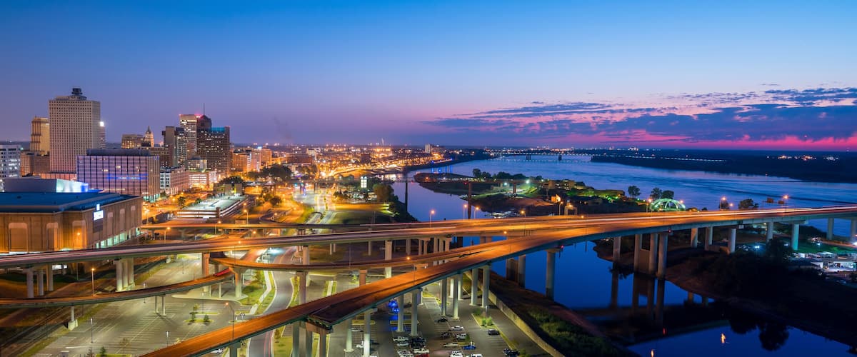 A cityscape at dusk with illuminated buildings, a river, and a bridge.