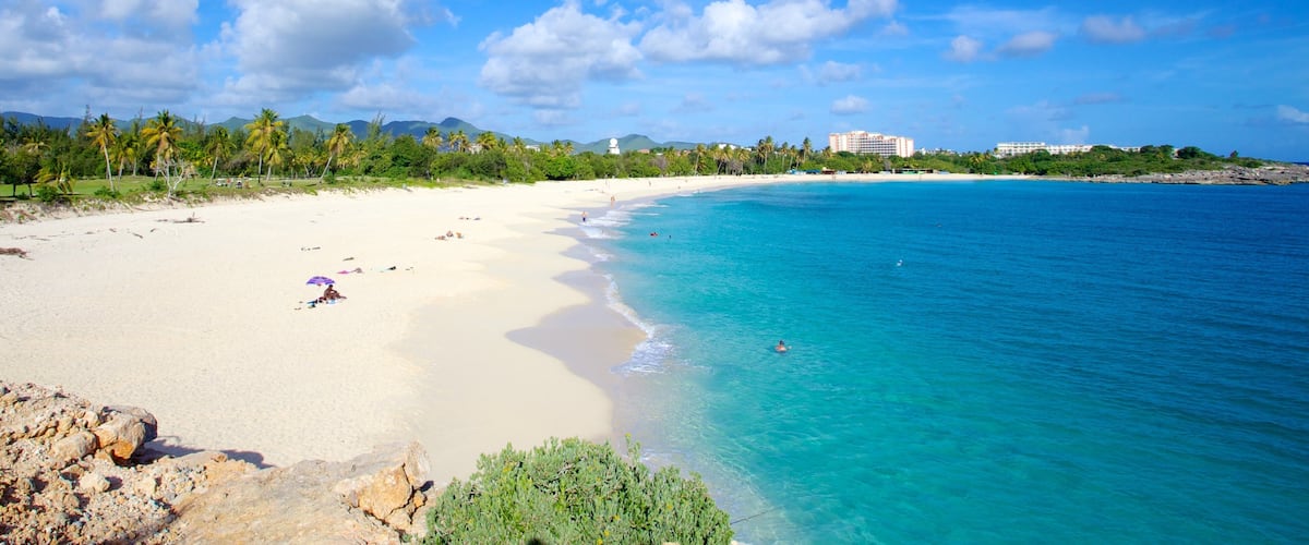 A beach with clear turquoise water, white sand, and palm trees. There are buildings in the background.