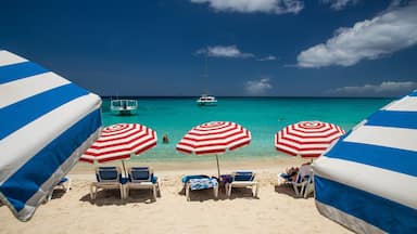 Une plage avec des parasols bleus et blancs à rayures, une eau turquoise cristalline et un voilier à l’horizon.