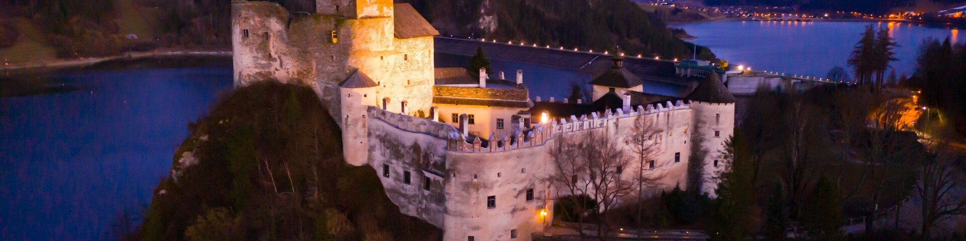 Un castillo en una colina con vista a un lago, rodeado de colinas boscosas.