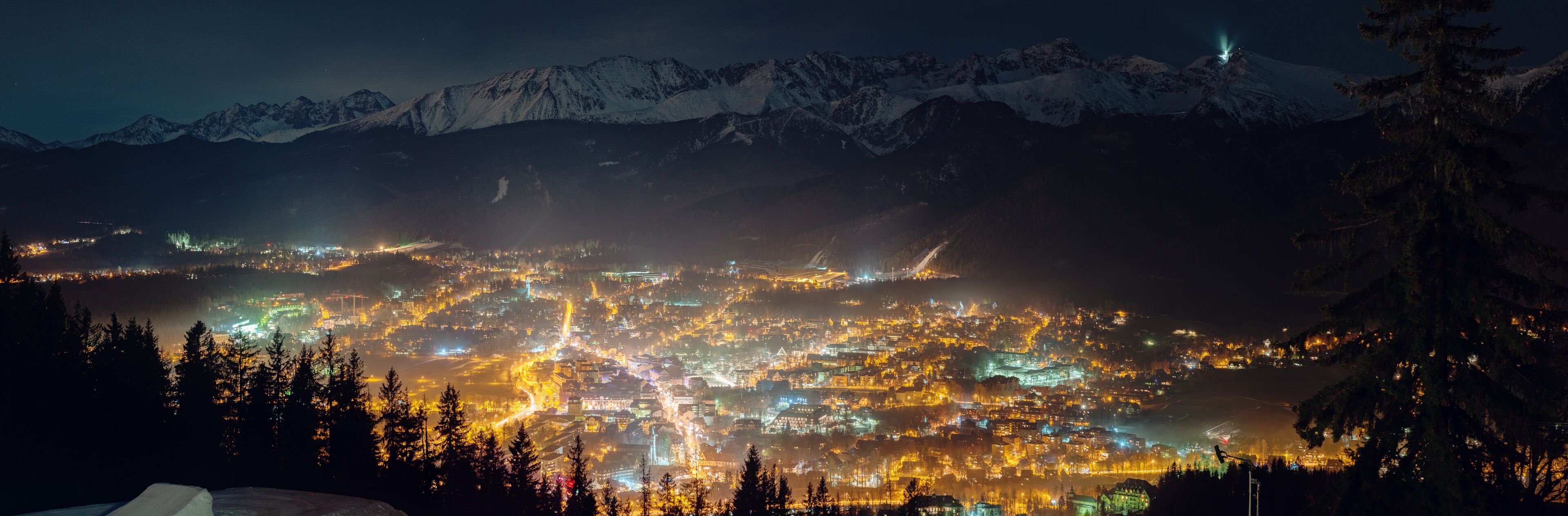 A nighttime view of a town nestled in a valley with snow-capped mountains in the background.