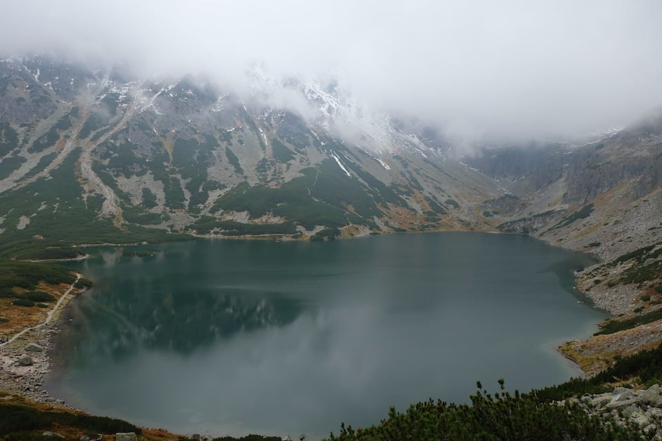 Ein See, umgeben von Bergen mit Schneeflecken und grüner Vegetation.