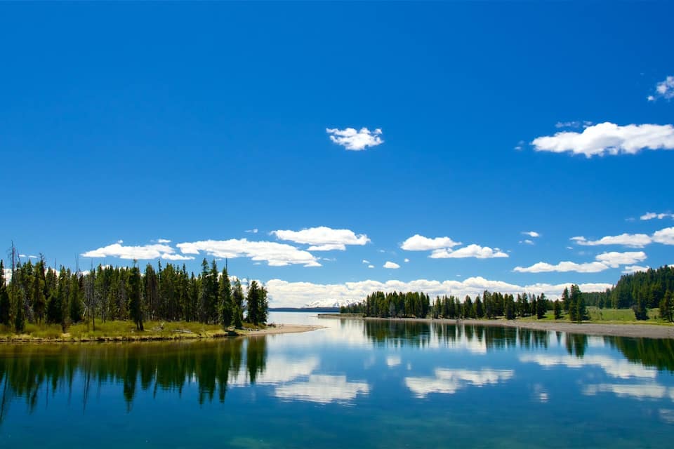 Um lago tranquilo com reflexos de árvores e céu, rodeado por florestas densas e uma cordilheira distante.