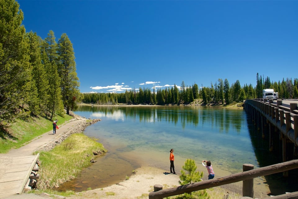 Um lago límpido com uma ponte de madeira, uma trilha de terra e uma área arborizada com montanhas ao fundo.
