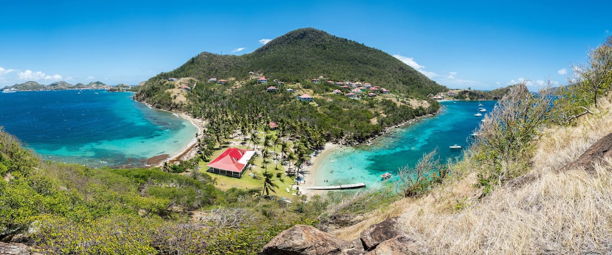 A coastal landscape with a mountain, clear blue waters, and a small settlement with red-roofed buildings.