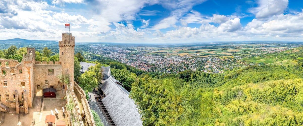 A historic castle with a flag, surrounded by lush greenery and a panoramic view of a town and distant mountains.