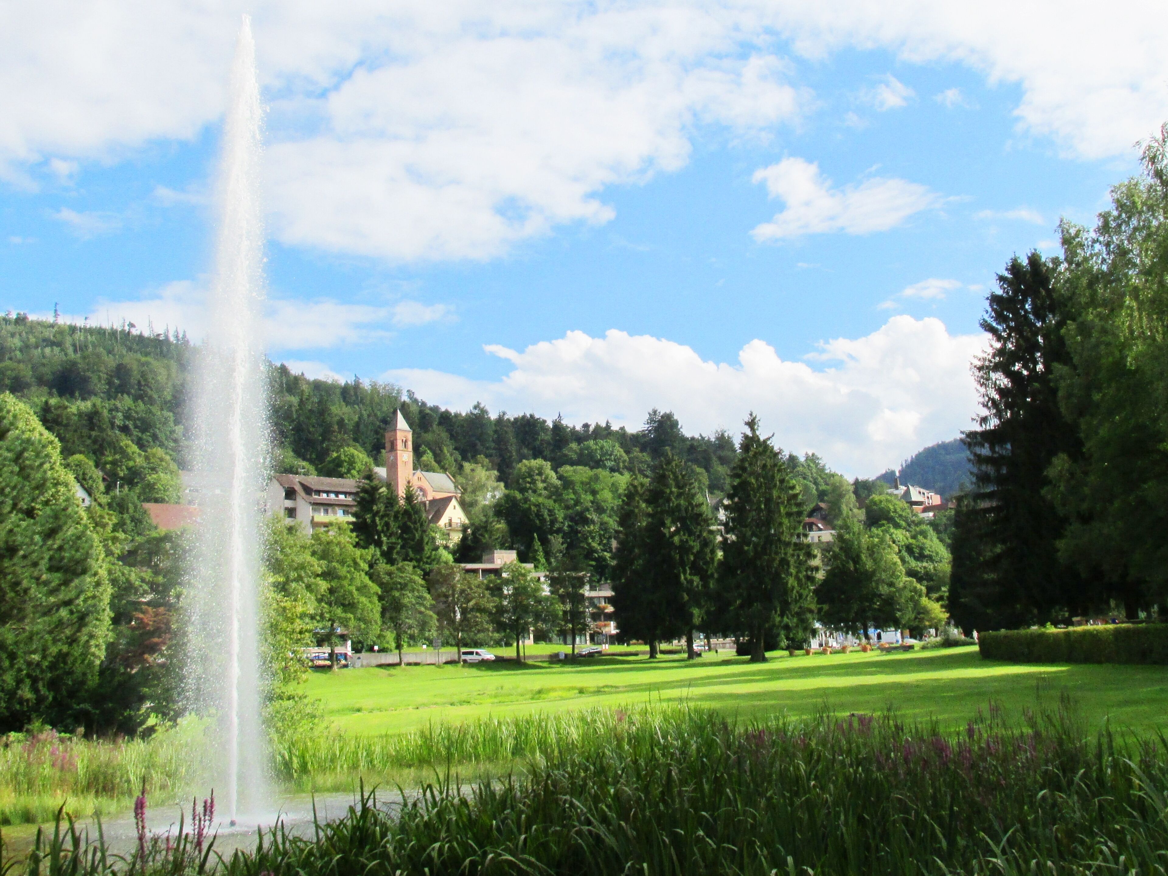 A fountain, a church, and a green park with trees and a hill in the background.