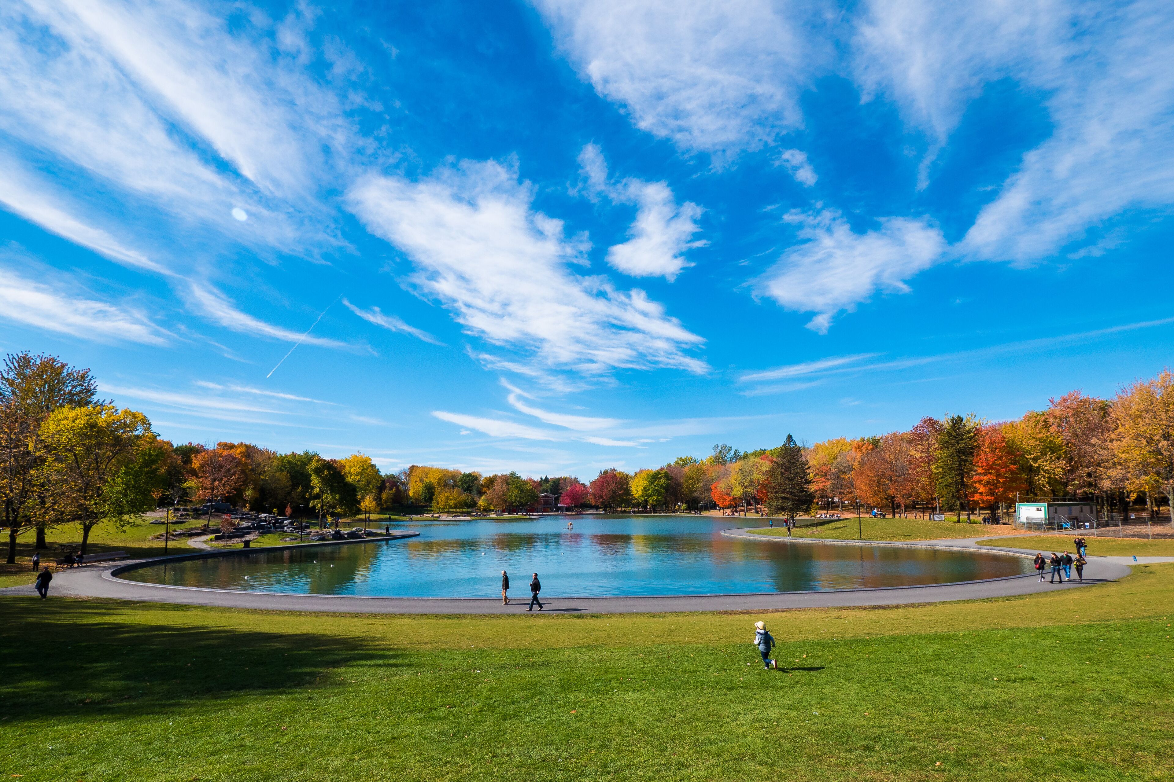 Beaver Lake on Mount Royal in Montreal, below a cloudy blue sky in autumn, with colorful maple trees and distant people around.
