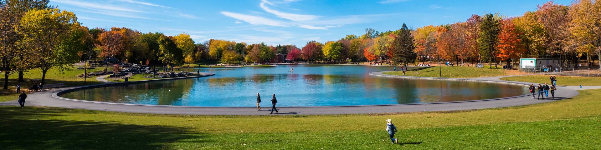 A park with a large pond, autumn-colored trees, and a clear blue sky.
