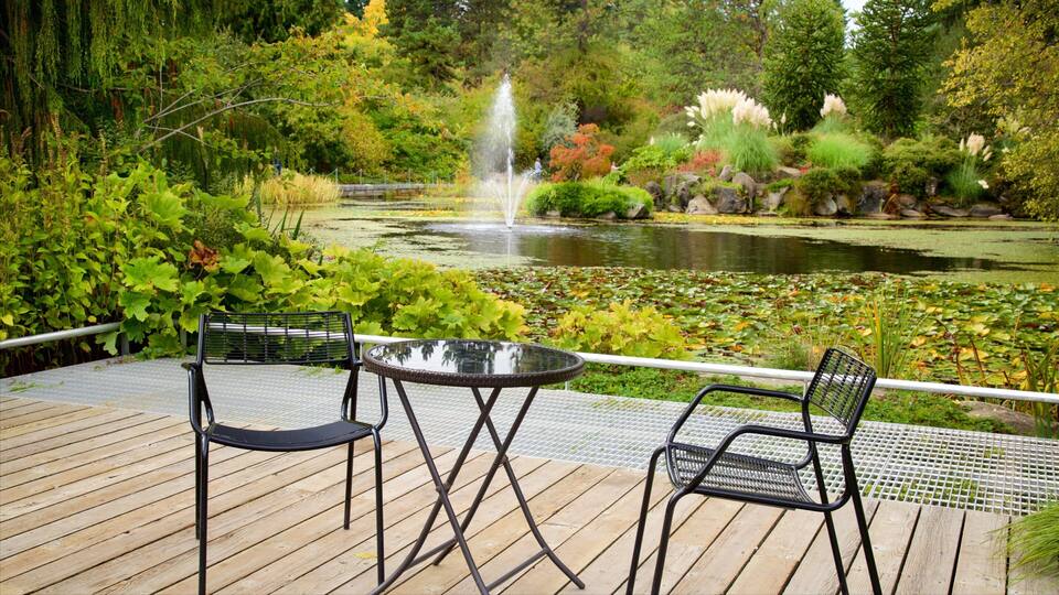 A wooden deck with two metal chairs and a table overlooking a pond with a fountain, surrounded by lush greenery and various plants.