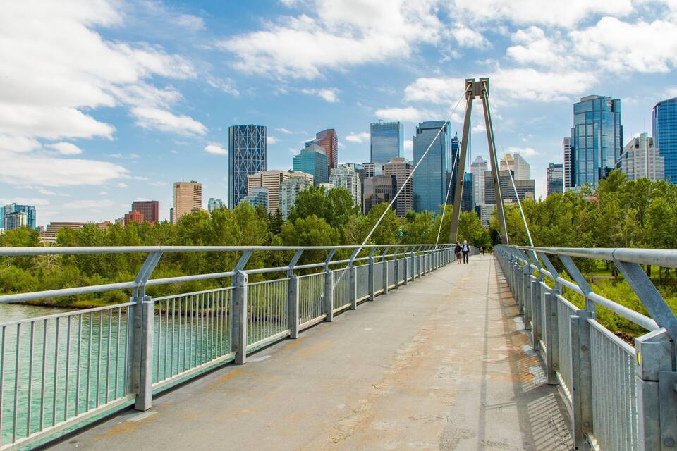 Un puente peatonal con barandillas metálicas, un horizonte urbano de fondo y una masa de agua a la izquierda.