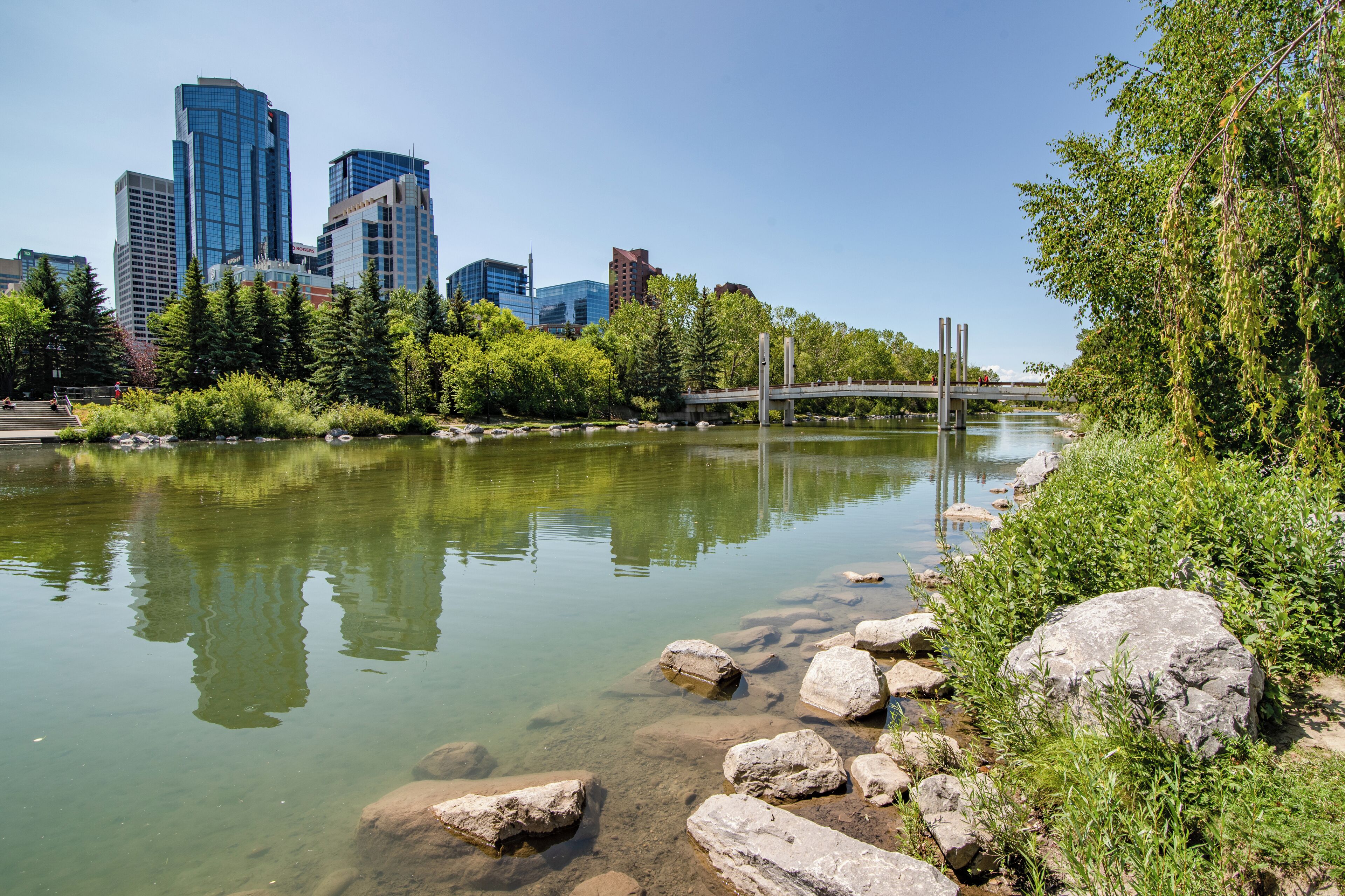 A river with clear water, surrounded by rocks and greenery, with a city skyline in the background.