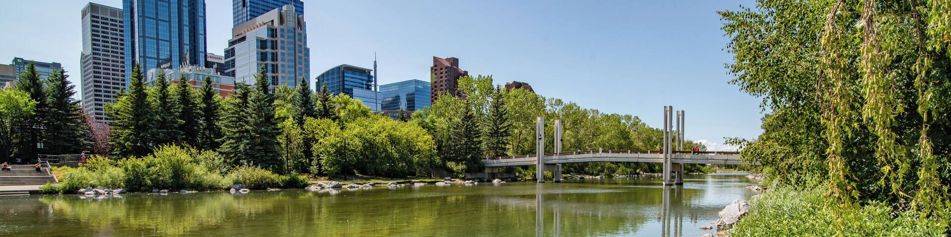 A river with clear water, surrounded by rocks and greenery, with a city skyline in the background.
