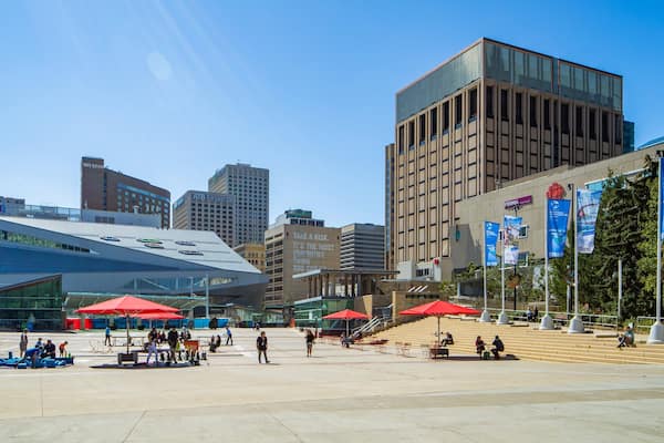 Uma praça urbana com edifícios modernos, guarda-sóis vermelhos e pessoas.