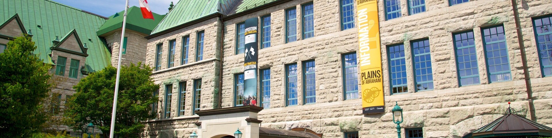 A historic stone building with a green roof, a Canadian flag, and banners advertising an exhibition.