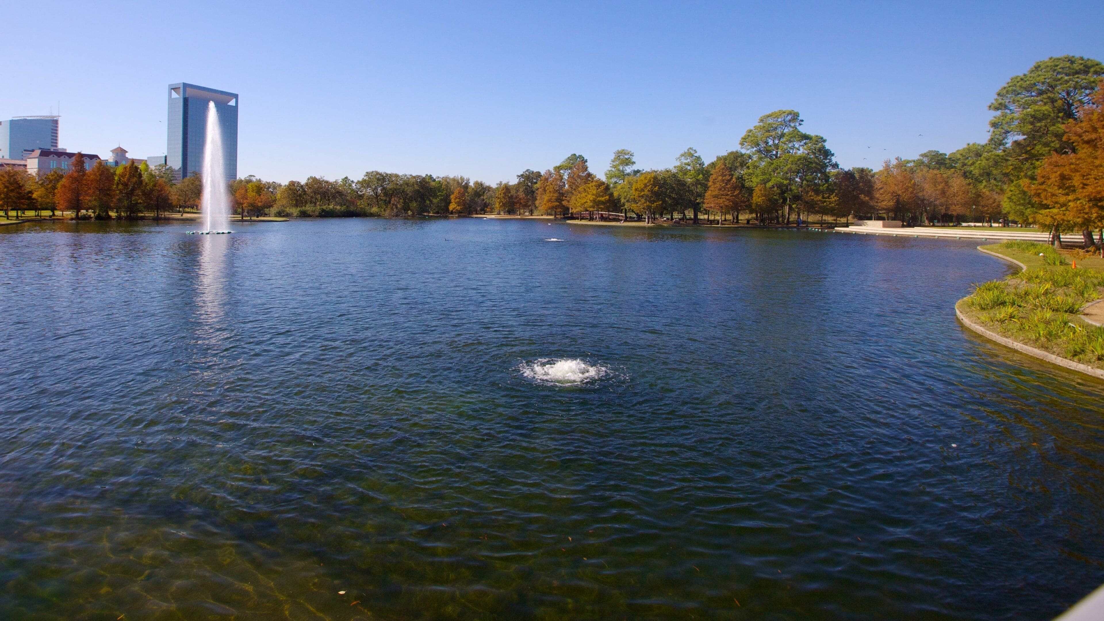 A lake with a fountain, surrounded by trees and a modern building in the background.