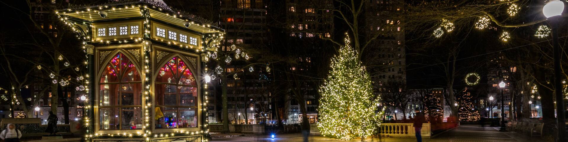 A brightly lit gazebo with intricate designs, a large Christmas tree adorned with lights, and a festive atmosphere in a city square at night.