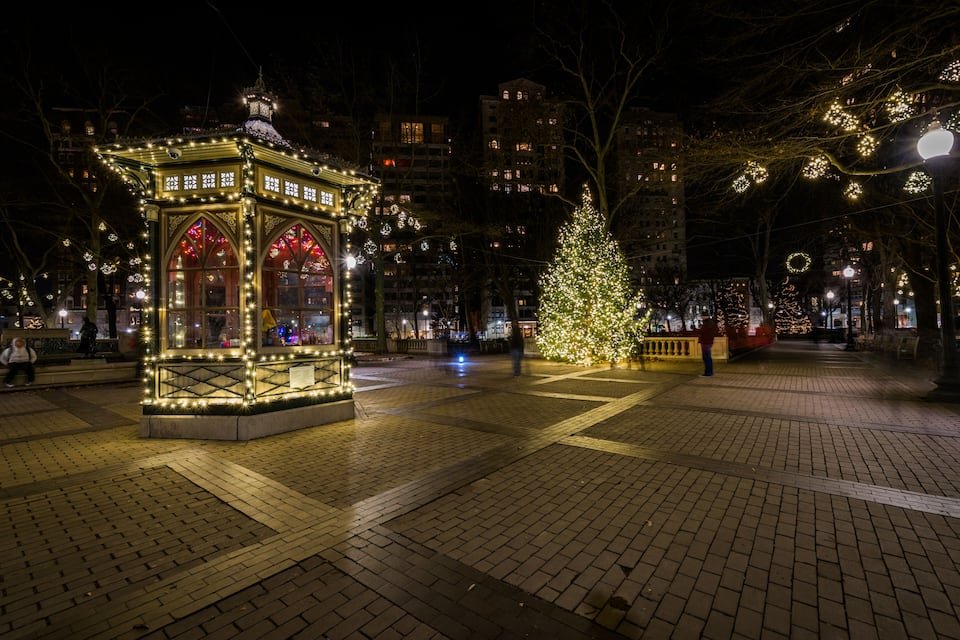 A brightly lit gazebo with intricate designs, a large Christmas tree adorned with lights, and a festive atmosphere in a city square at night.