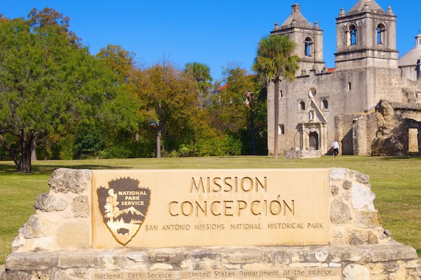 En historisk bygning med et klokketårn, omgivet af træer og et skilt for Mission Concepcion National Historical Park.