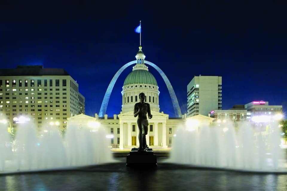 Paesaggio urbano notturno con una statua in primo piano, un grande edificio a cupola sullo sfondo e una fontana con getti d'acqua.