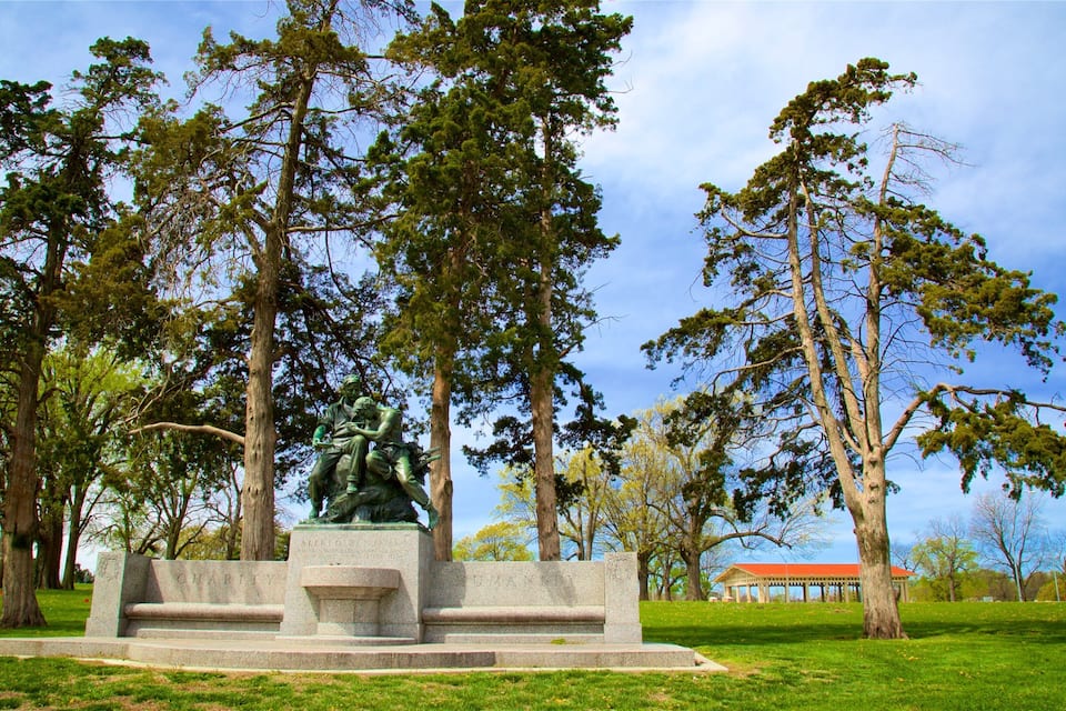 A park with a statue of two figures, surrounded by tall trees and a pavilion in the background.