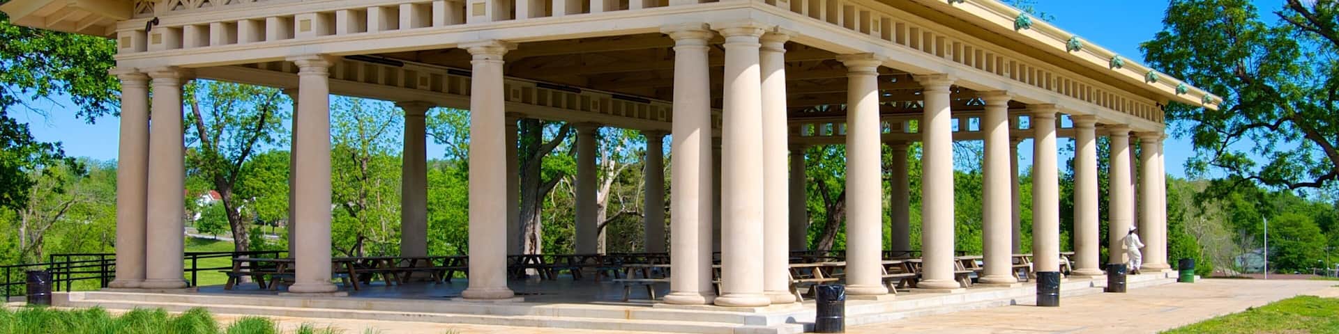 A classical pavilion with a central open space surrounded by columns, set in a park with grass and trees.
