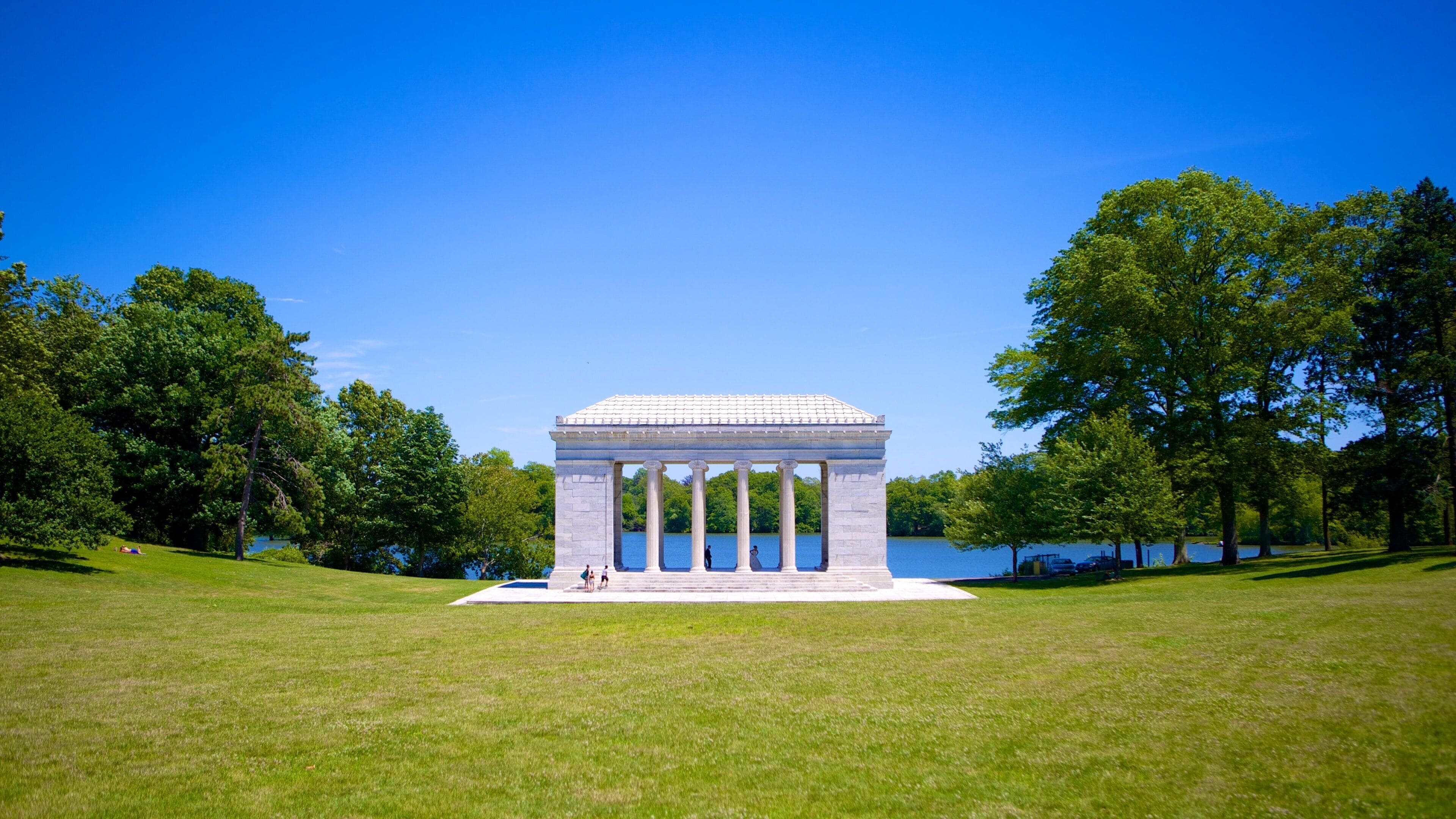 A neoclassical monument with columns, surrounded by a grassy field and trees, with a lake in the background.