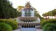 A tiered water fountain with a palm-like top, surrounded by well-manicured hedges and trees.