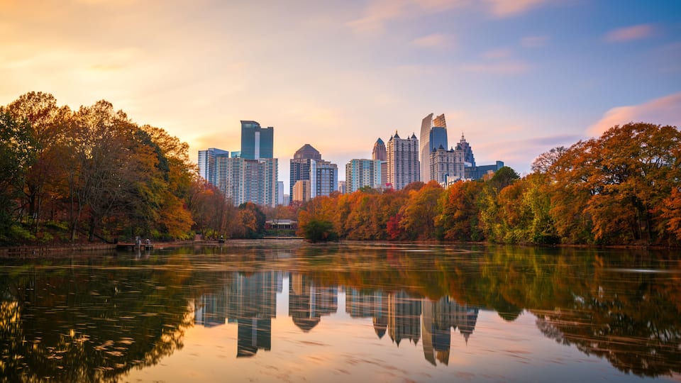 Ein Stadtpanorama spiegelt sich in einem ruhigen See, umgeben von herbstlich gefärbten Bäumen.