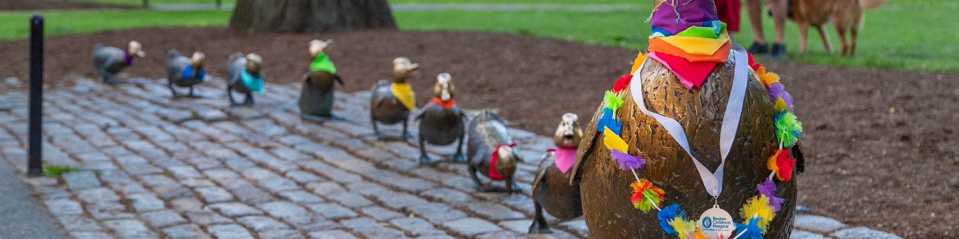 Fileira de esculturas de bronze de pássaros com decorações coloridas, em um caminho de paralelepípedos em um parque.