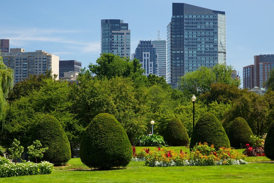 A park with manicured hedges, flowering plants, and a city skyline in the background.